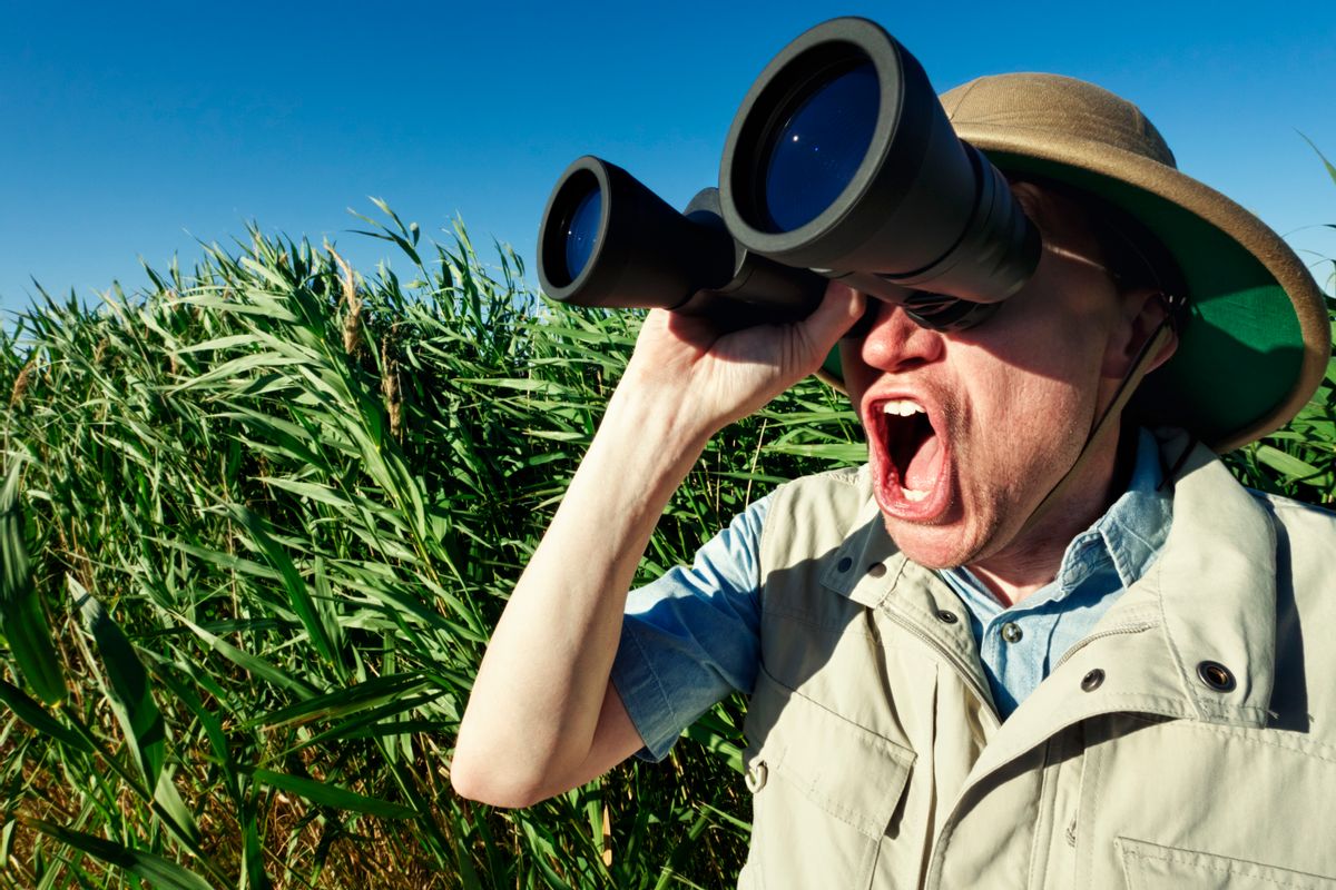 Photo of a bird watcher standing in tall grass, looking through binoculars with an amazed expression. ( jhorrocks/Getty Images)