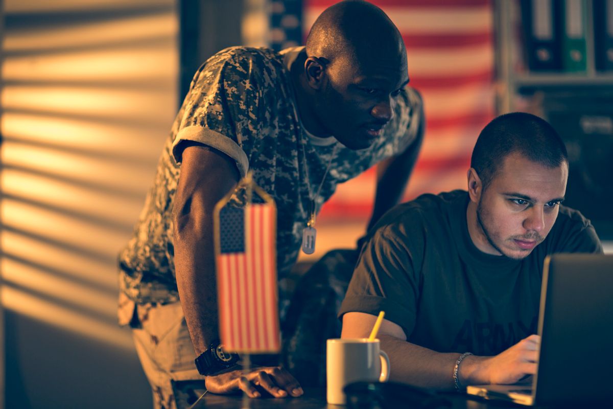 Photo of two young american soldiers looking at a laptop on the desk together. One is sitting and one is standing, On the desk is also a small flag and a coffee mug. In the background is a bookshelf and on the wall is hanging an american flag. The soldiers look tired and a little bit sad. From the left a dim light is sipping threw the window. (Getty Images/
Geber86) (Getty Images/Geber86)
