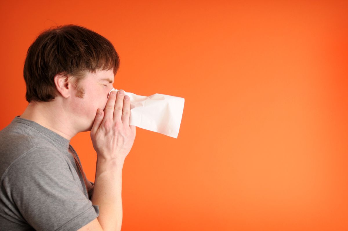 Color photo of a man sneezing or blowing his nose into a facial tissue in front of an orange background. Room for text on right. (Getty Images)