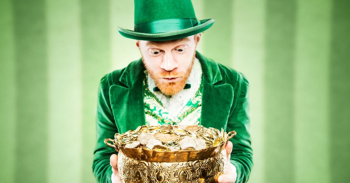 A stereotypical Irish character all ready for Saint Patricks day.  He holds up a pot of gold looking at it with wide excited eyes.  Geen striped wallpaper wall behind him.  Horizontal image with copy space. (RyanJLane/Getty Images)