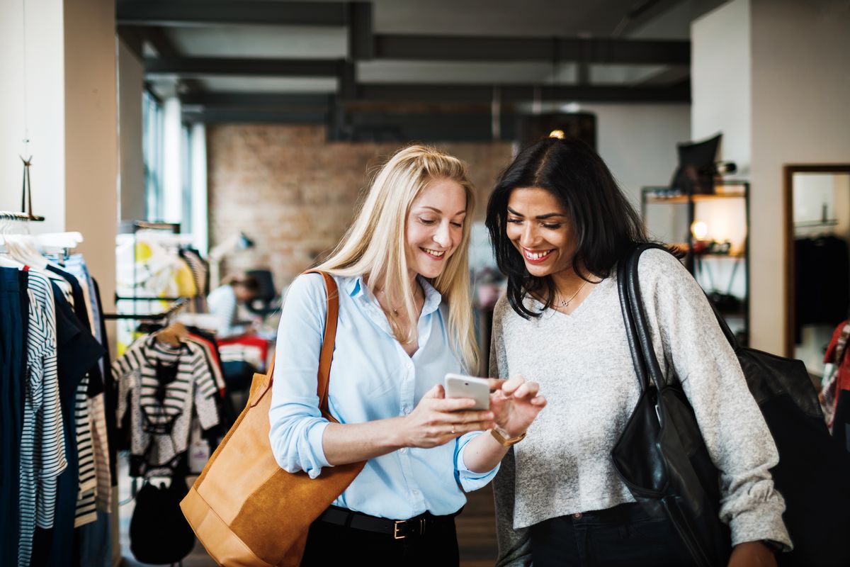 Two women smiling and looking at a smartphone while out shopping for the day together. (Getty Images/Stock photo)