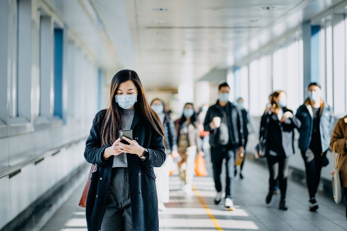 Asian woman with protective face mask using smartphone while commuting in the urban bridge in city against crowd of people (Getty Images/Stock photo)