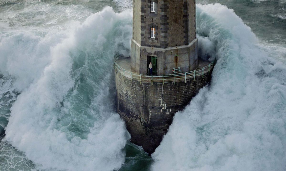 A photograph shows a big wave hitting a lighthouse while a lighthouse keeper stands in the doorway and the picture is from 1989 at the La Jument Lighthouse in France. (Jean Guichard)