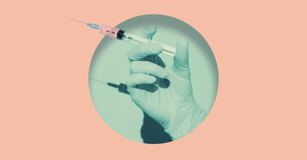 Doctor's hand holding syringe of Coronavirus vaccine placed inside round hole in pink paper.Digital composite"n (Getty Images)