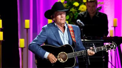 A white man wearing a blue suit and black cowboy hat stands before a microphone.