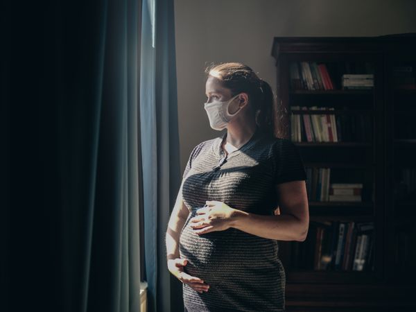 Pregnant woman with face mask standing in front of window. (Getty Images)