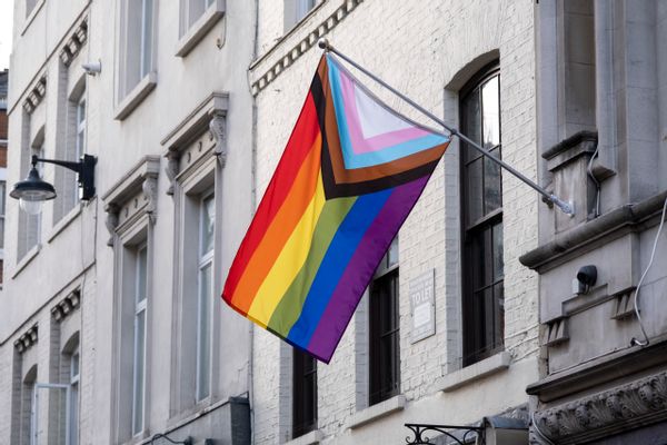 A flag with a rainbow, as well as black, brown, light blue, light pink, and white stripes in the shape of a triangle on the side, hang from a white building.
