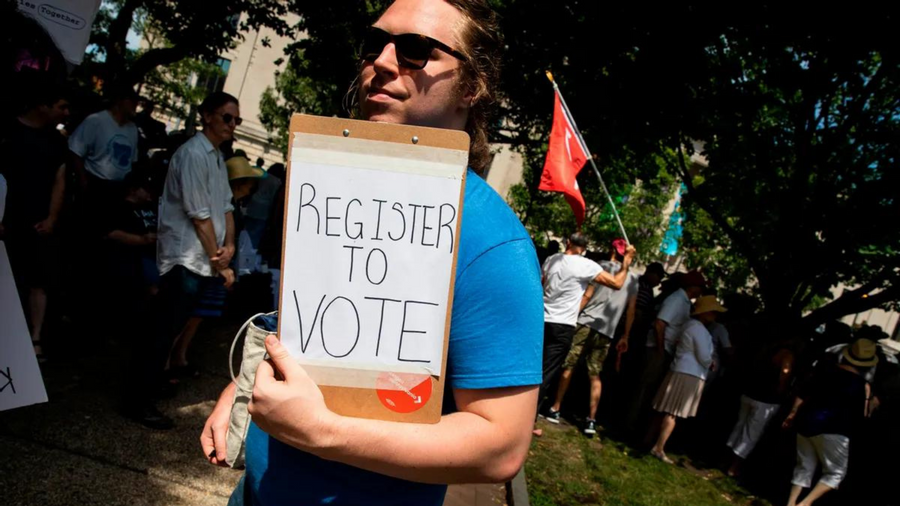 A white person wearing sunglasses holds a clipboard that says REGISTER TO VOTE on a piece of paper. A crowd of people, some holding flags, can be seen in the background.