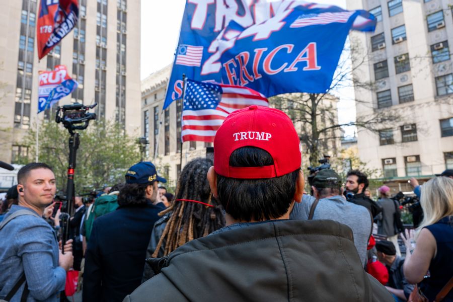 A person wearing a TRUMP hat stands near a crowd that holds flags that show support for former President Donald Trump.